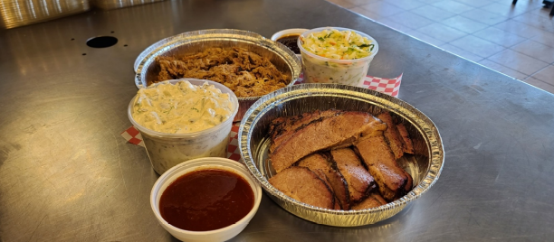 A catering spread displays foil containers of pulled meat, sides, and sauces.